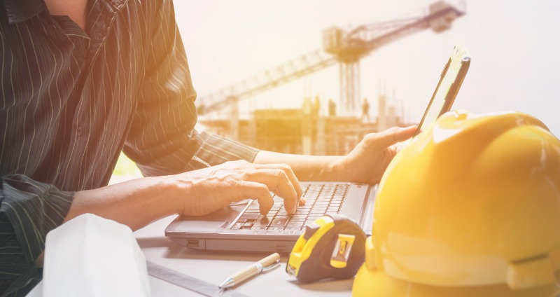 Man on site using his rugged laptop while sitting at a desk with a crane in the background.